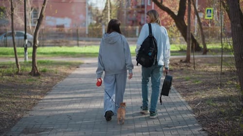 Two Friends Leaving Park with Small Dog White Male and Asian Female Backpacks Briefcase Leash
