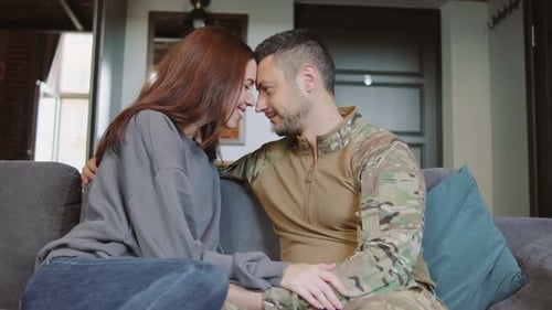 A Man in Military Uniform and a Woman Sit Close on a Couch at Home Exchanging Smiles and Touching