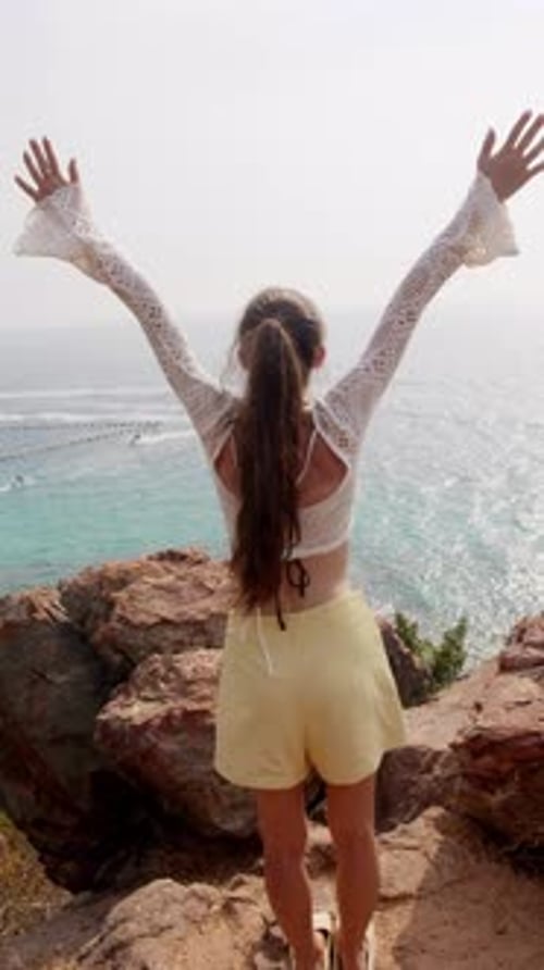 Une jeune beauté fait face à l'océan en étendant les bras au bord de la falaise en admirant les vagues bleues et les feuilles vertes de l'horizon