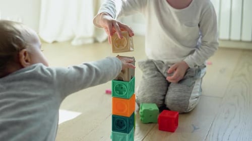 Two Childs Building Tower of Block Toys Sitting on Floor in Sunny Bedroom