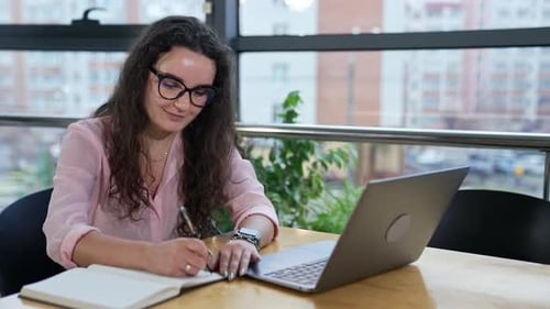 Smiling curly-haired lady taking notes sitting in front of laptop.