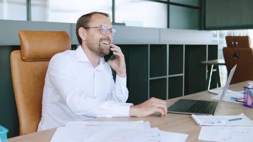 Businessman Talking on Phone While Working with Laptop at Desk in Modern Office During Daytime