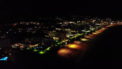 Aerial Over Chairs And Parasols Lit By Street Lighting Over Beach At Night In Majorca, Spain.