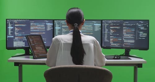 Asiatische Entwicklerin arbeitet mit Tablet, während sie Code mit mehreren Bildschirmen auf grünem Bildschirm schreibt