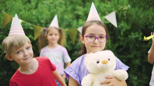 Uma menina em uma camiseta colorida sorrindo com um ursinho de pelúcia em uma festa de aniversário ao ar livre com Happy