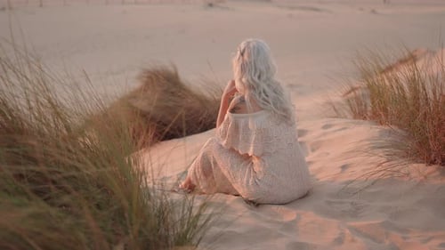 Serene Woman Sits on Sand Dune Contemplating Sunset Over Ocean Waves in Golden Hour Light