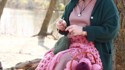 A girl knits clothes from wool while sitting on the roots of a tree on the river bank