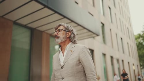 Mature businessman wearing gray jacket walks down the street past modern buildings