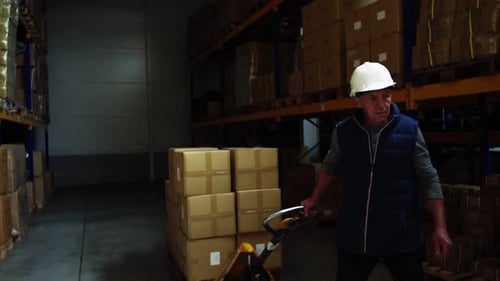 Senior warehouse worker pulling a pallet truck through a busy distribution center