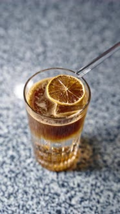 Ice cube in the glass with brown beverage. Long metal tongs putting the slice of dried orange on top