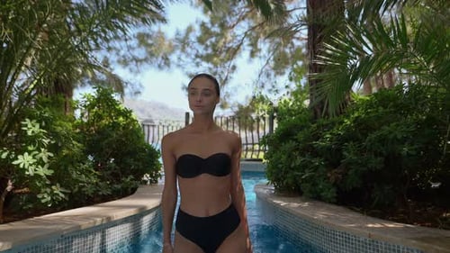 Young woman in black bikini standing in a shallow pool surrounded by lush greenery and palm trees