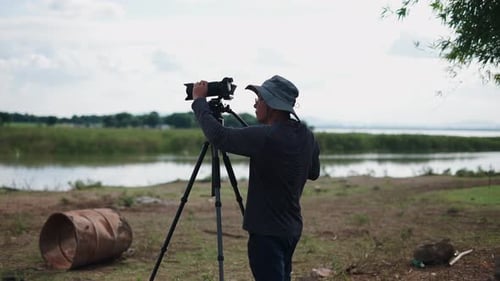 a Photographer Adjusts His Camera on a Tripod on the Dirty Plain Near the River in Thailand