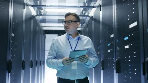 IT Technician Walks Through Rows of Server Racks in Data Center. Simultaneously He Works on a Table