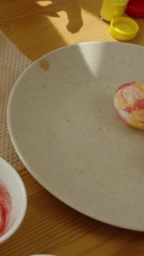 Vidéo verticale. Rouler à la main un œuf de Pâques sur un tapis peint, avec des bols de colorant et des œufs blancs dessus