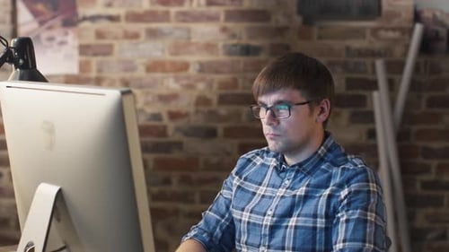 Young man in glasses is sitting in front of a monitor in a loft and working.