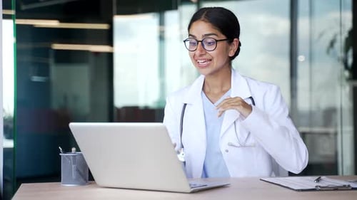 Young female doctor in white coat and glasses talking on video call using laptop in modern hospital