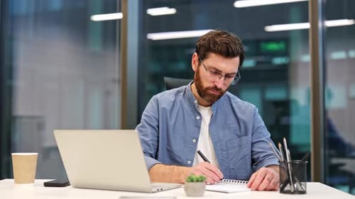 Businessman Working at Laptop in Office Writing in Notebook