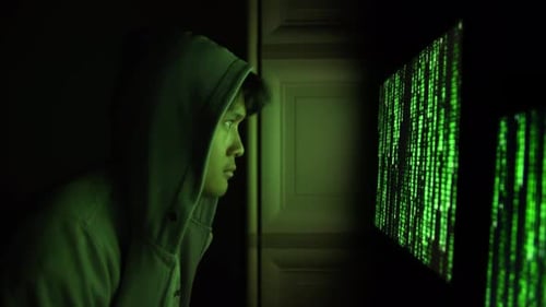 Un hacker cagoulé fait face à un écran de code numérique dans un cyberenvironnement sombre