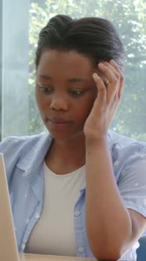 Vertical video: Reacting to laptop alert, woman touching head at desk by window, blue shirt, ring