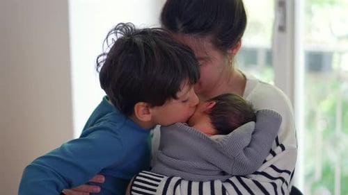 Sweet Boy Kisses Mother Holding Newborn Baby Indoors
