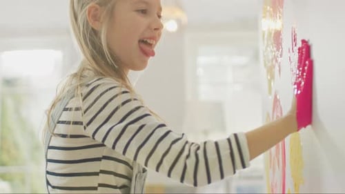 Happy Little Girl with Hands Dipped in Yellow and Pink Paint Puts a Colorful Handprint on the Wall.