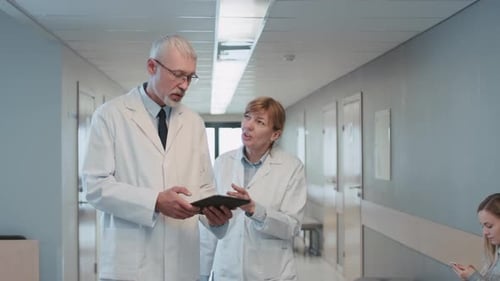 In the Hospital Two Doctors Walking Through the Hospital Hallway, They Talk and Share Tablet Comput