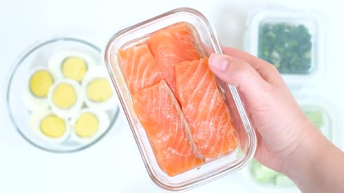 Mano femenina que sostiene una lonchera de cristal con salmón sobre fondo blanco.