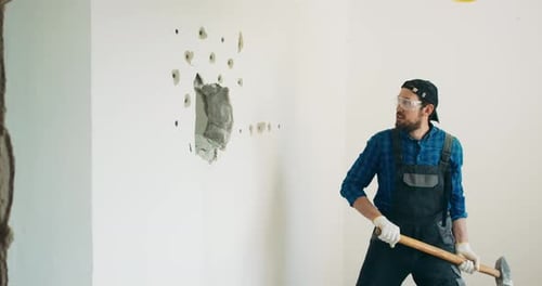 A Smiling Guy Demolishes a Wall in a House Using a Construction Hammer on a Long Stick