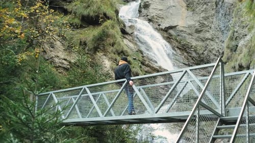 Man in Jeans Jacket Walking on Bridge Over Wild Mountain Waterfall