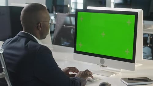 Business African Man Working on Desktop Computer with Green Screen Mock Up Display in Busy Creative