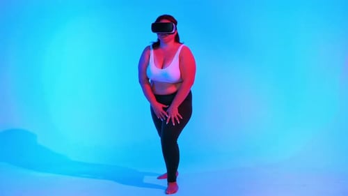 Woman with overweight in a tracksuit and VR headset posing in a studio with blue background and red