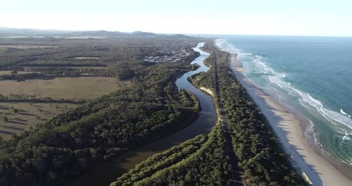 Aerial shot of a river at sunset with mountains in the background and the ocean with breaking waves