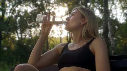 Young Woman Sits on Bench in Park Forest in Morning After Jogging and Drinks Water