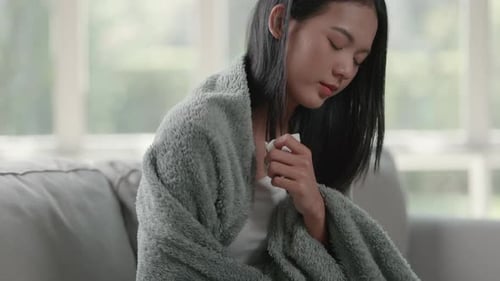 Unhappy sad Asian woman in cloak suffering from fever and flu on sofa