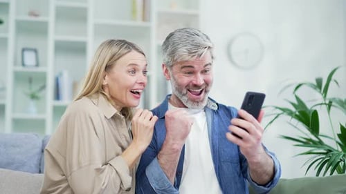 Close up of amazed happy married couple checking smartphone, reading message while sitting at home.