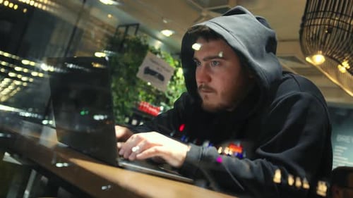 A man is programming, typing a code on a laptop at night in an institution, possibly a hacker