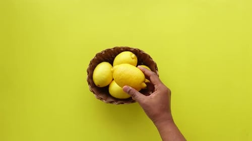 A Hand Reaching Out for Fresh Juicy Lemons in a Decorative Basket on a Bright Yellow Background