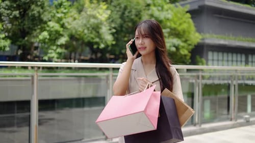 Young woman smiles while talking on phone near a shopping mall. She holds several shopping bags