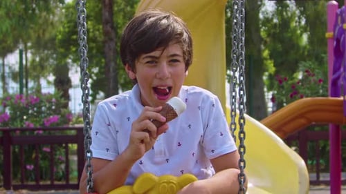 Teenage Male Eating Ice Cream While Swinging On A Swing In The Playground