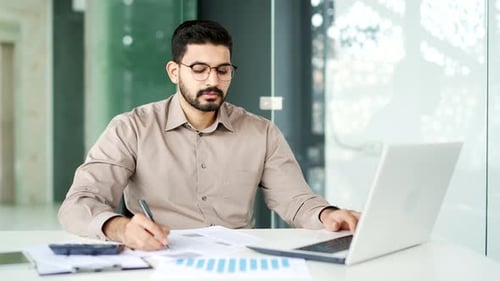 Busy financier is doing paperwork using a laptop computer while sitting at the workplace. Confident