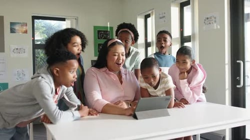 Female teacher with six children tapping tablet on white table in classroom sharing content