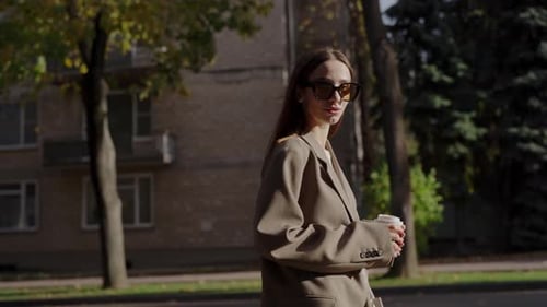 Young Woman in Sunglasses Walks Past City Buildings with Paper Cup of Coffee
