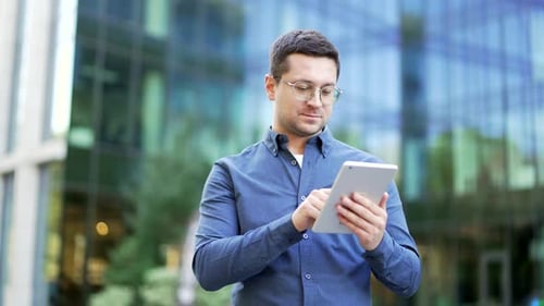 Selbstbewusster Geschäftsmann benutzt ein digitales Tablet, während er auf der Straße in der Nähe eines modernen Büros steht