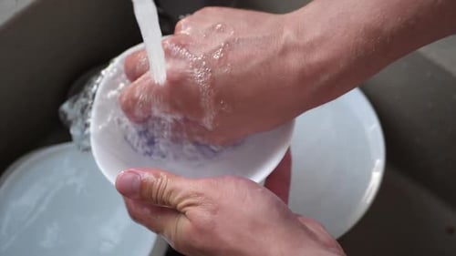 A man washes dirty dishes with a sponge under the pressure of water.