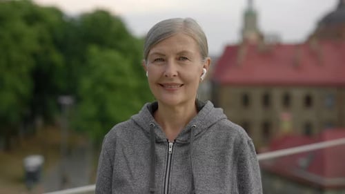 Close Up Portrait of the Smiled Mature Woman Wearing Earphones in Sporty Clothes Standing in Tje