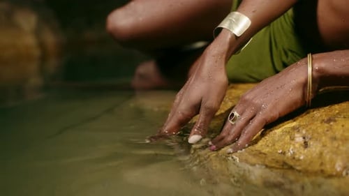 Nonbinary Black Person Hand in Brass Rings with Gems Golden Bracelets Touches Water Lying on Big