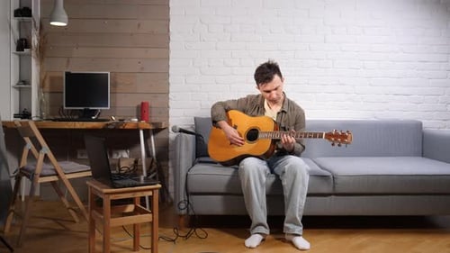 Jeune homme se relaxant sur un canapé tout en jouant de la guitare acoustique s'adonnant à un passe-temps créatif et