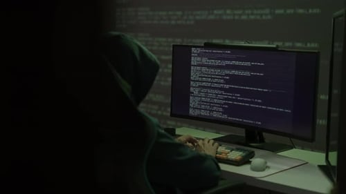 Hooded Figure Typing Code at Computer in Dark Room