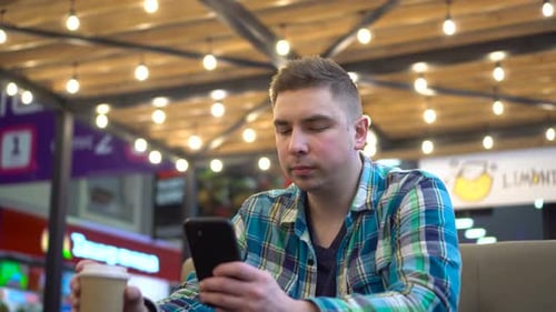 A Young Man Sits in a Cafe with a Phone in His Hands and Drinks Coffee