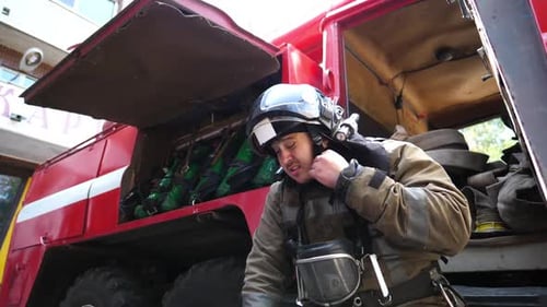 Männlicher Feuerwehrmann nimmt den Helm vor dem Hintergrund eines roten Lastwagens ab, junger Feuerwehrmann entfernt